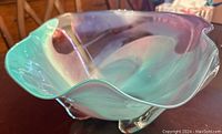 Front angle view of the signed hand-blown glass bowl showing its swirl of green and purple colors and wavy rim.