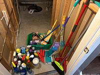Wide view of cleaning supplies in basement with brooms, brushes, chemical bottles, and cleaning tools arranged on floor.