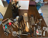 Top view of large collection of cooking utensils including spatulas, wooden spoons, ladles, scoops, a potato ricer, can opener and other utensils spread on a table beside the two black caddies.