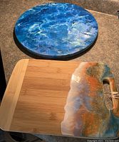 Photo showing the 14-inch round Lazy Susan with a glossy blue, white, and green fluid acrylic paint design and the bamboo charcuterie board with resin paint accent.