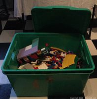 Top down view of green plastic storage bin filled with assorted LEGO bricks and pieces including colored plates and curved track pieces.