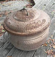 Metal fire pot with lid placed on outdoor wooden deck, showing rusty exterior and circular handles.
