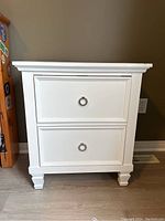 Front view of white wooden night stand with two drawers and silver ring handles.