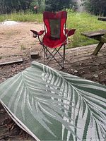 Red folding camping chair on wooden deck. Pale green outdoor rug partially rolled up on ground nearby with white leaf pattern.