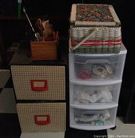 Vintage woven sewing basket on top of 3-drawer plastic organizer and 2-drawer cardboard storage unit with wooden box of scissors next to them.