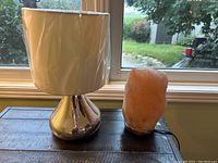 Photo of silver table lamp with white shade and pink-orange Himalayan salt lamp side by side on a wooden surface near a window.