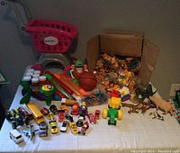 Overview of the toy collection including miniature cars, animals, bubble wands, shopping cart, and construction vehicles in a box.