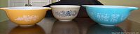 Front view of three vintage Pyrex mixing bowls in a row showing different colors and designs: yellow Butterfly Gold, white and blue Homestead, and turquoise Amish Butterprint
