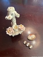 Front angle photo of the white spaghetti poodle figurine with pink and yellow flowers and the set of one large and three small cameo pins laid on a wooden surface.
