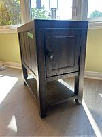 View of dark wooden side table from an angle showing the small door on one end, lower shelf, and textured wood finish.