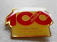 Gold-tone enamel pin celebrating Coca-Cola's 100 year centennial with bright red and gold colors.