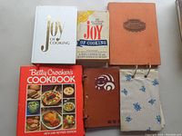 View of six cookbooks including two Joy of Cooking editions, Betty Crocker's Cookbook, leather-bound handwritten book, orange embossed book, and fabric-covered book.