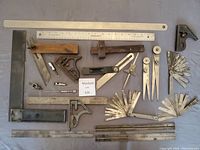 Collection of metal and wooden machine shop measurement and marking tools including squares, rulers, bevel gauges, marking gauge, dividers, calipers, depth gauge and feeler gauges.