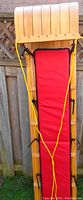 Front view of vintage wooden toboggan showing slatted wood, curved front, red padded seat and yellow ropes.