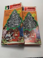 Two unopened boxes of vintage Double-Glo tinsel garland with packaging showing Christmas tree shape and silver garland inside cutout windows.