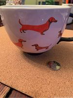 Side view of large white coffee mug with printed dachshund dogs in red and brown. Coin placed beside mug to show scale.