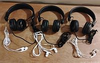 Three black JLAB headphones with padded ear cups and attached wires with 3.5mm jacks, laid on wooden surface with three pairs of wired white earbuds.