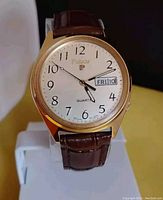 Front view of the watch displayed on a stand showing the linen dial, gold-tone case, and brown leather strap.
