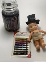 Photo of the lot showing a sealed pack of Singer fabric crayons with six colors, a Yankee Candle jar candle with dark purple wax, and a 1959 New Years baby doll with a black top hat labeled '1959' and wrapped in beige cloth.