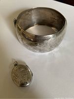 Photo showing the wide sterling silver bracelet alongside the engraved oval locket, both tarnished and aged.