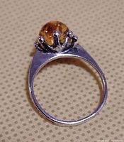 Top view of sterling silver ring showcasing crown-like prong setting holding round amber-colored stone with faceted details, on beige textured background.