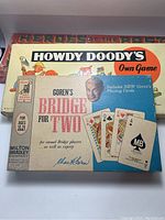 Photo of three vintage game boxes stacked: Howdy Doody's Own Game on top, Goren's Bridge for Two in front, and Heroes of the Past puzzle partially visible below.