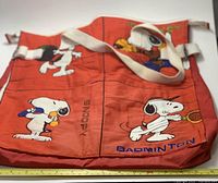 Front view of red fabric sports bag with Snoopy characters playing badminton and visible fabric wear.