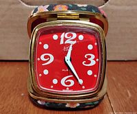 Front view of the square red-faced clock with white numbers 12, 3, 6, and 9, and brass-tone frame, housed in floral fabric covered case.