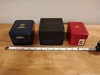 Three vintage watch boxes from left to right: blue Waltham, black Seiko, red Jules Jurgensen, with measuring tape showing length approximately 10 inches total.