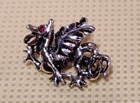 Close-up photo of a small Welsh dragon brooch made of silver or silver-plated material with red stones for eyes, displayed on a beige textured background.