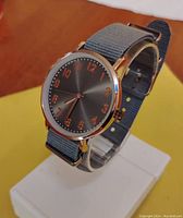 Photo showing watch face with grey dial, rose gold numerals, and grey NATO strap on display stand.