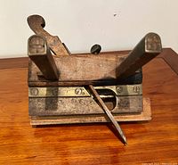 Front view showing the plane's wooden body, double vertical handles with capped ends, brass accents, and angled metal blade for planing wood.