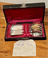 Full view of the set of 12 coffee spoons neatly arranged inside a fitted red velvet-lined black presentation box with latch closure, including a handwritten note saying 'WMF Kaffee Löffel 90 x'