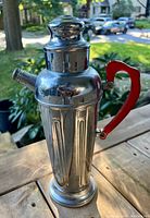 Front and perspective view of the mid century cocktail shaker showing its metal body with vertical grooves and red bakelite handle.