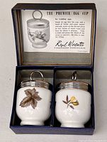 Two porcelain egg coddler cups with floral design and metal lids in original box showing instruction card inside the lid.