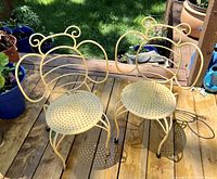 Pair of yellow painted vintage cast iron kids chairs with bumble bee design backs, perforated round seats, and four curved legs shown outdoors on wood decking with plants in background.