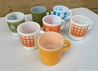 Photo shows seven vintage Fire King coffee cups in various mid century colors and patterns on a white table.