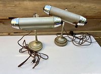 Two vintage library lamps side by side on a white surface, showing cylindrical metal shades with silver ends and brown cords.