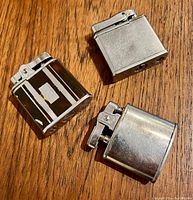 Three vintage metal lighters displayed on wooden surface showing different shapes and finishes.