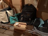 Lot of bags on metal rack showing black Lucas backpack, beige Lacoste toiletry bag, teal Candi sack, and other bags