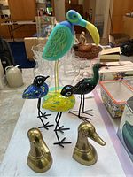Photo showing two brass duck busts in front with three colorful wooden and gourd beach birds behind, exhibiting painted details and metal legs, with various home items around.