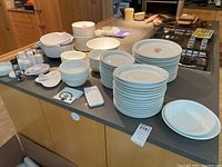 Overview photo showing stacks of white ceramic plates, Corelle bowls, white salt and pepper shakers, plastic mixing bowls, and other kitchen accessories on kitchen island