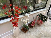 Four faux orchid floral arrangements placed by window, including tall white vase with red flowers and smaller vases with white and pink orchids.