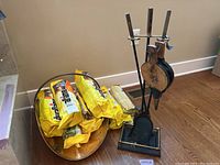 Wide view of fireplace tool set next to brass tone log holder filled with yellow Duraflame log packages on hardwood floor.