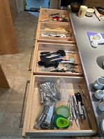 Four open wooden drawers with assorted kitchen utensils and flatware inside, seen in natural light in kitchen area.