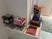 Photo showing stacked board games, puzzles, and toy boxes including Scattergories, Light Brite, dominoes, and wooden train whistle plus small American flags.