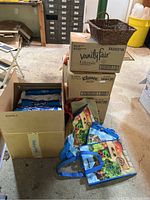 Photo showing three stacked boxes: Vanity Fair napkins on top, Kleenex tissues in middle, Charmin toilet paper at bottom. Wicker basket placed on top of boxes. Several reusable shopping bags on the floor next to the boxes.
