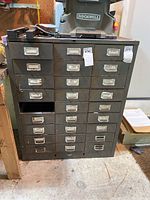 Front view of 27-drawer metal cabinet showing all drawers with labels. One drawer missing at bottom right.