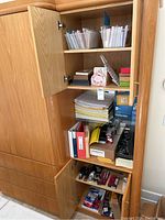 Wooden cabinet storing office supplies including binders, lined paper pads, staplers, and boxes.
