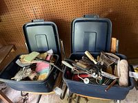 Two blue plastic storage bins filled with assorted painting supplies including brushes, rollers, scrapers, and rags, with lids open to show contents.